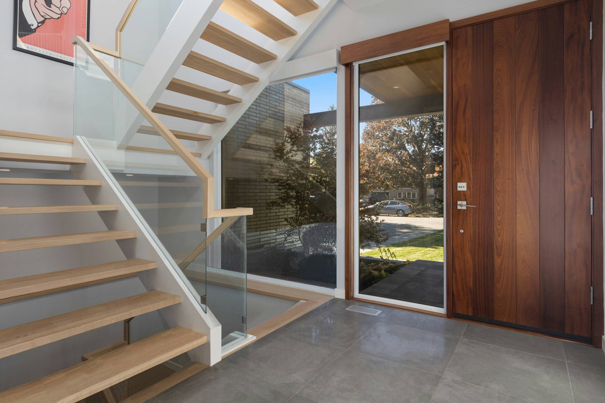 Open-riser floating stair with thick light-oak treads and a frameless glass guard, rising beside a tall window in a modern entry